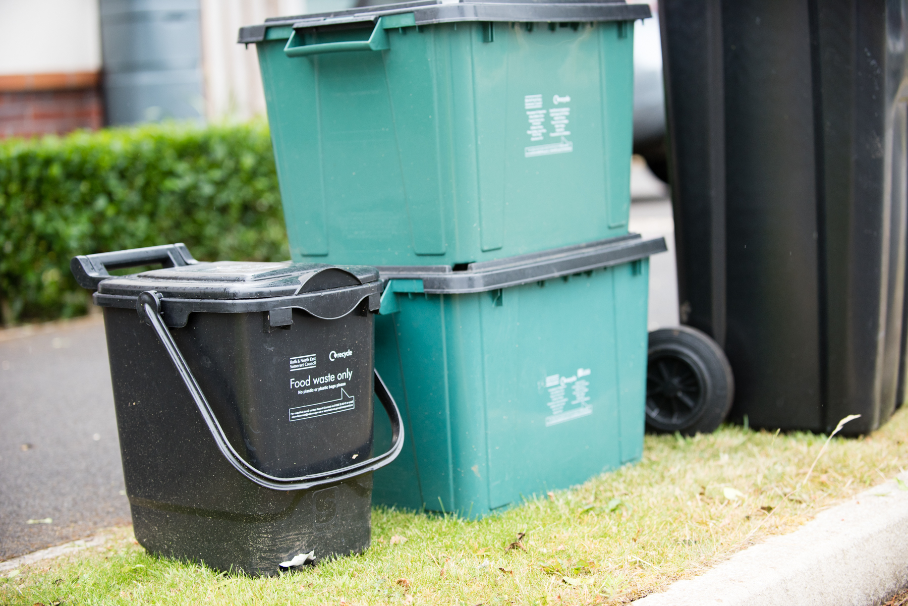 a photo of a black food waste recyling bin alongside two green recycling boxes and a black waste bin. All on a kerbside
