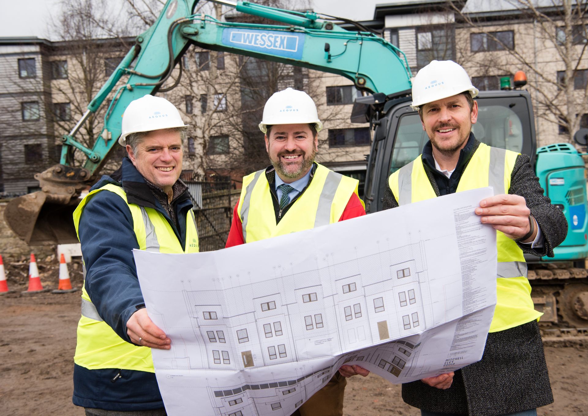 Three men wearing white hard hats and yellow high visibility jackets looking at a plan of the development on site with a digger behind them