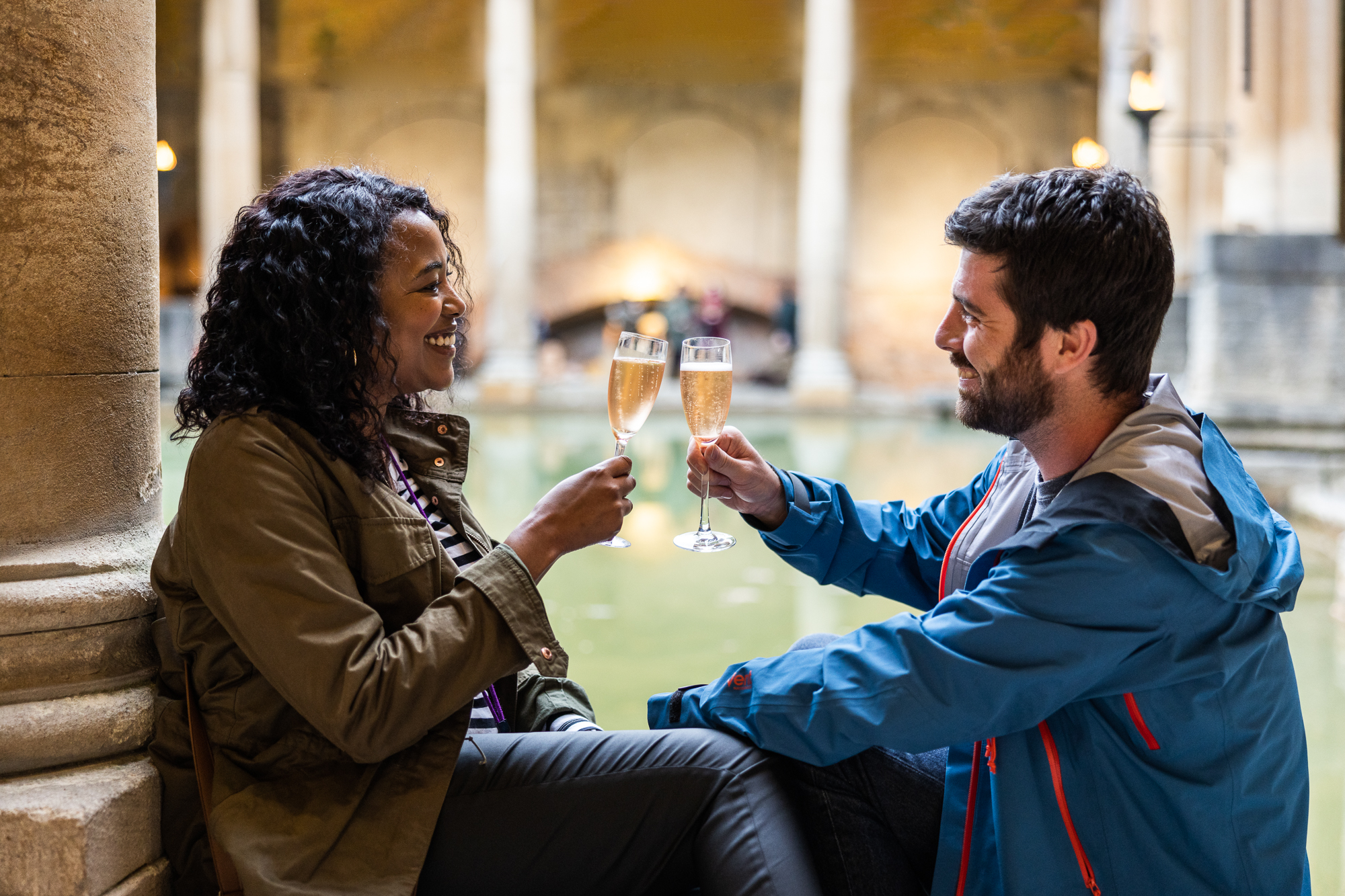 A woman and man toasting with champagne glasses beside the Great Bath