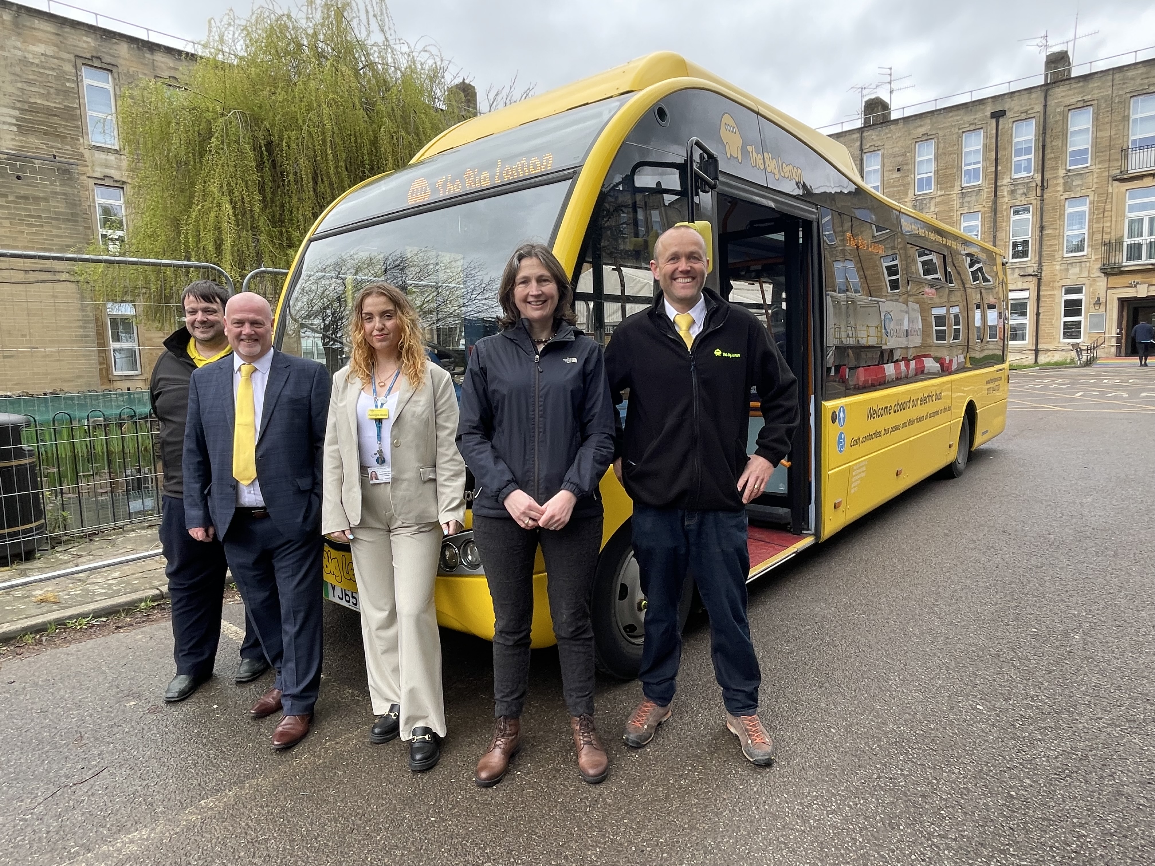 In front of a yellow bus at the Royal United Hospital: Photo caption (left to right): Tom Druitt, CEO at The Big Lemon; Councillor Sarah Warren; Georgia-Rose Gleeson, Sustainability Officer at The Royal United Hospital; Colin Morris, General Manager at The Big Lemon; Jason Freeman, Operations Manager Bristol & The West at The Big Lemon