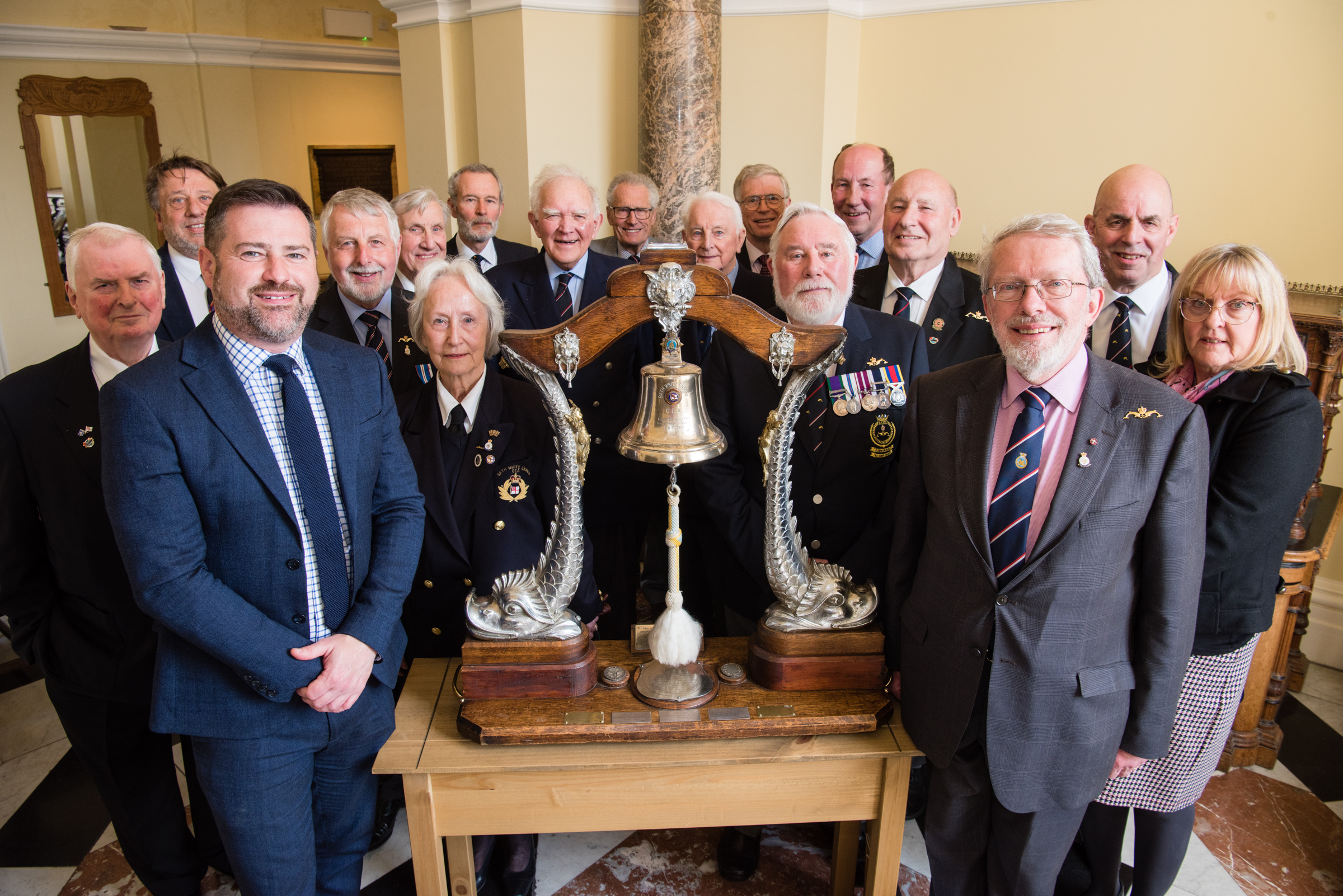 A group of people gather around a table on which is the historic Dolphin Bell, a bell mounted on a wooden frame with a silver dolphin either side of it.