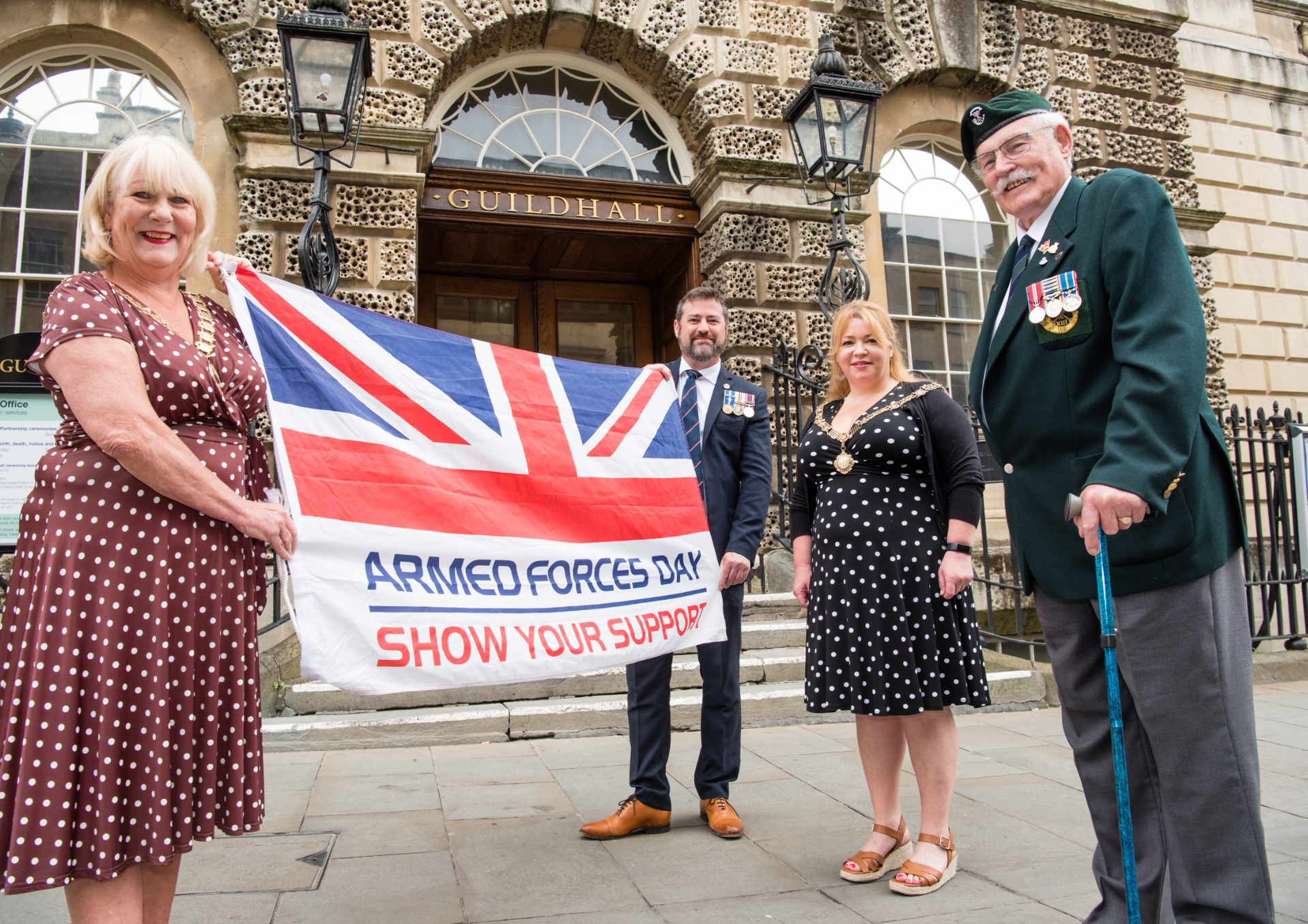 a photo of a woman and a man holding an Armed Forces Day flag with another woman and an older man in with medals on his chest outside the entrance to a building