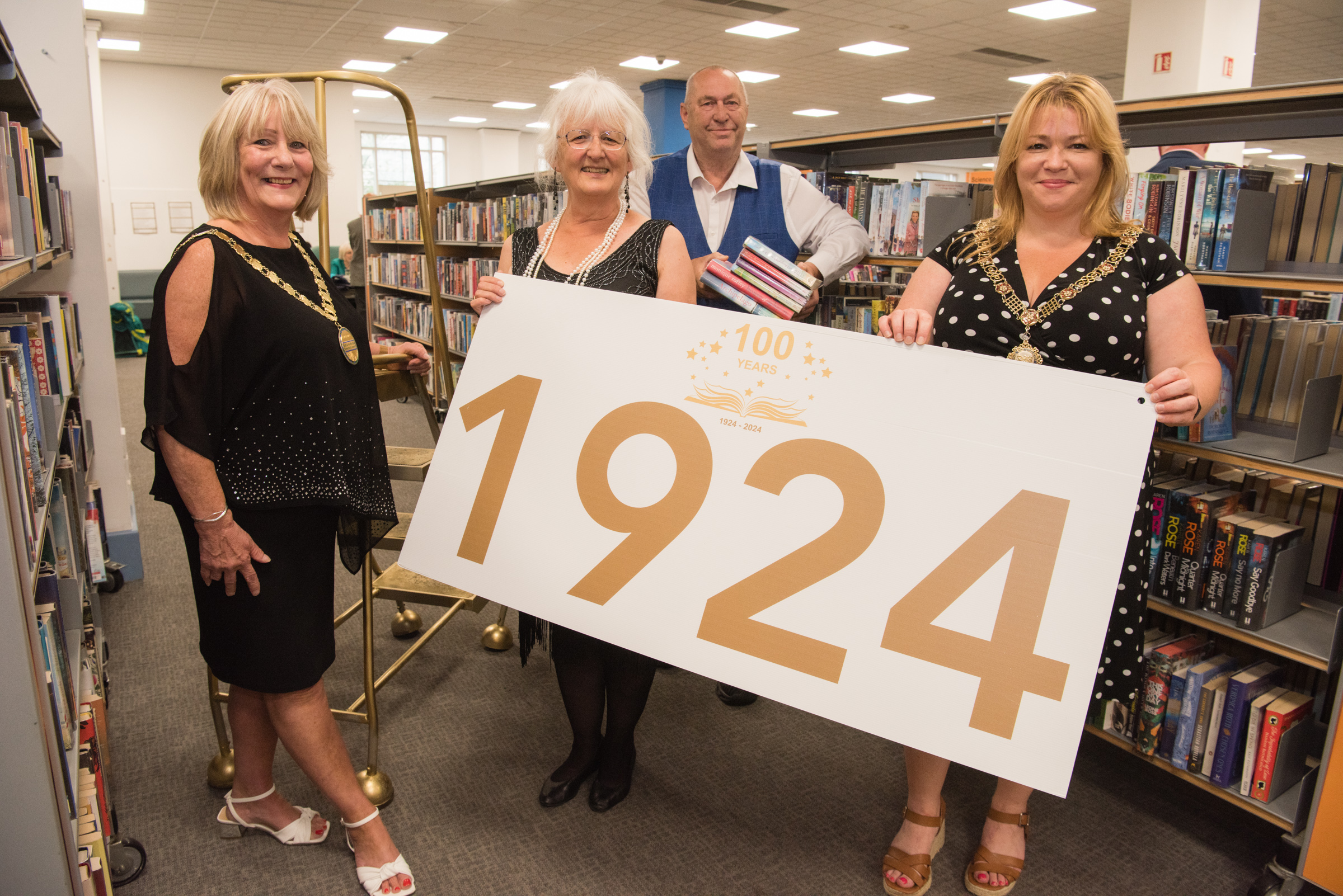 Four people hold a sign saying 1924. They stand next to bookshelves at Bath Central Library. In the background is a set of library steps painted gold.