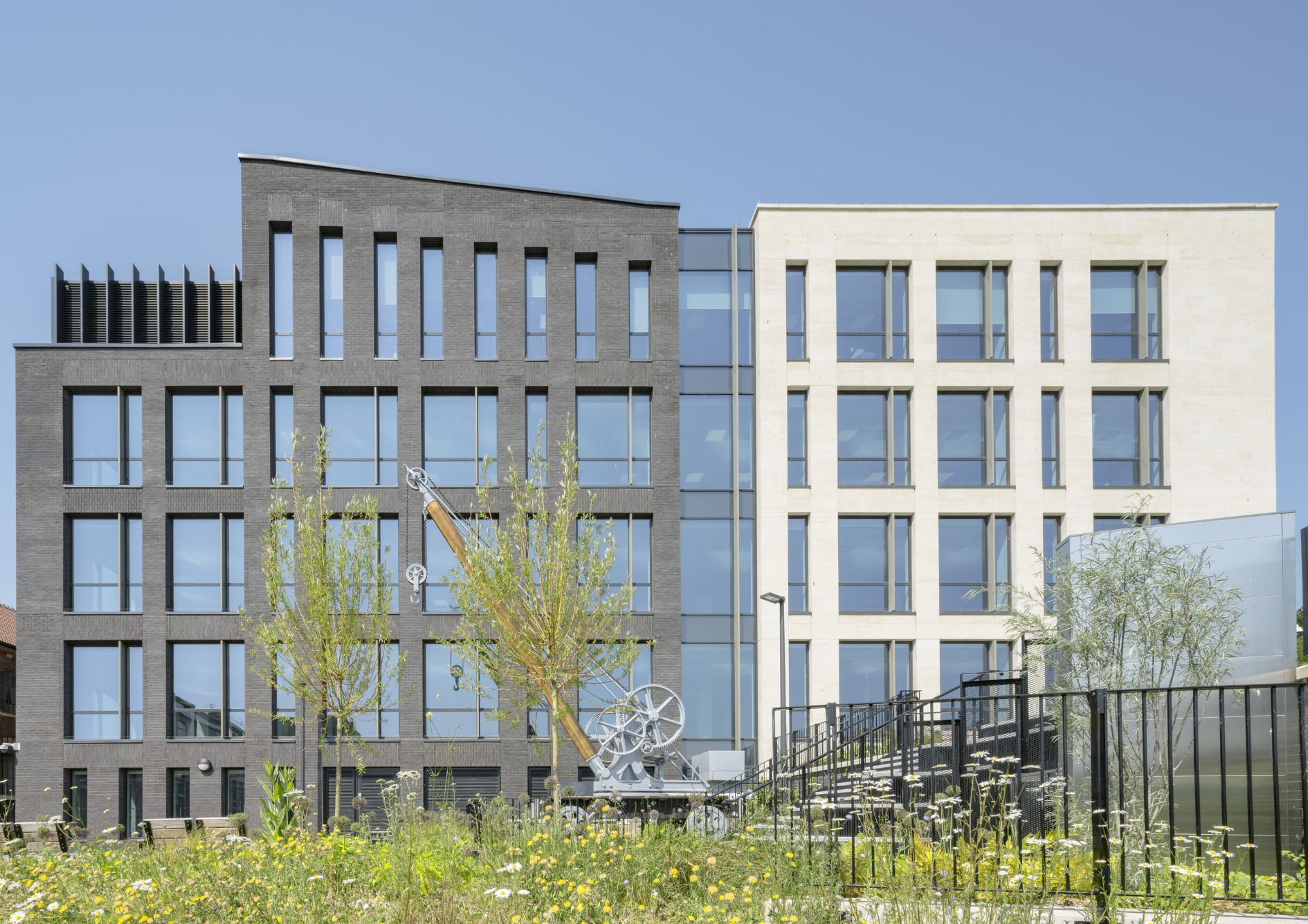 photo of the outside of an office block half grey and half white. In front are wild borders, a tree and an old crane.