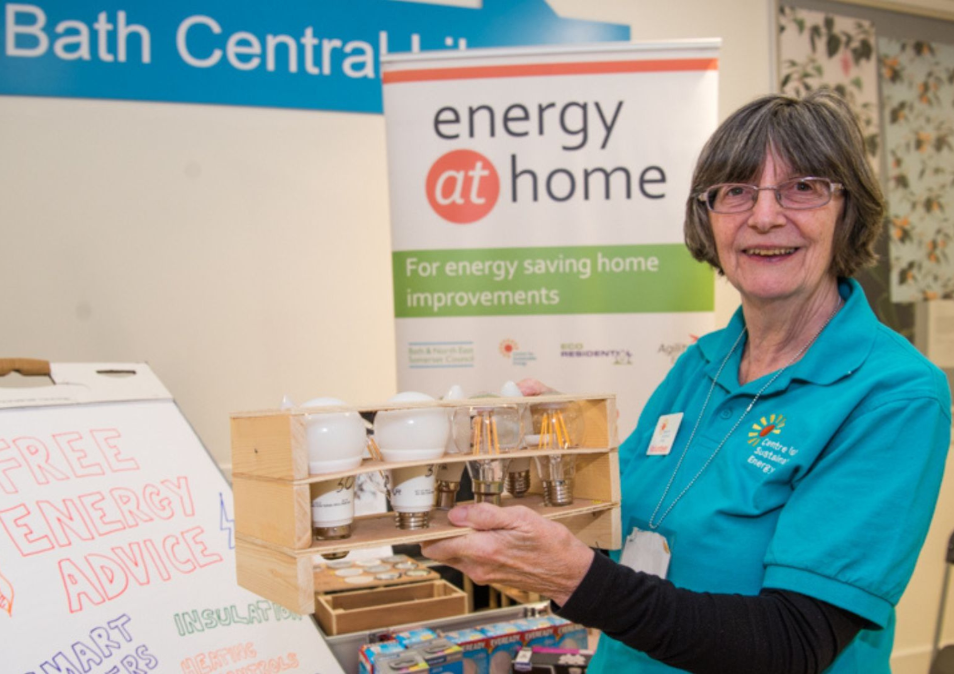 a lady holding a display of light bulbs in front of a stall and banner advertising energy at home