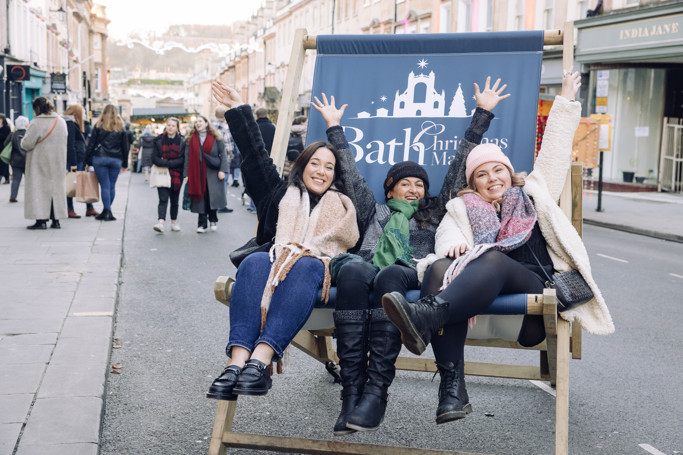 Three people sit in a giant Bath Christmas Market deckchair in Milsom Street in Bath. They hold their hands up in celebration. In the background are pedestrians and shop buildings either side.