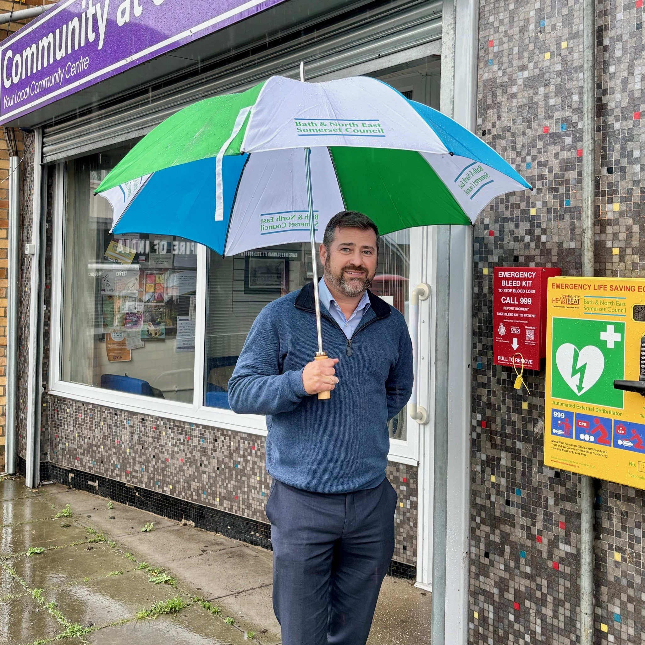 Councillor Kevin Guy stands holding an umbrella next to a bleed kit fixed on a wall outside the Community at 67 shopfront