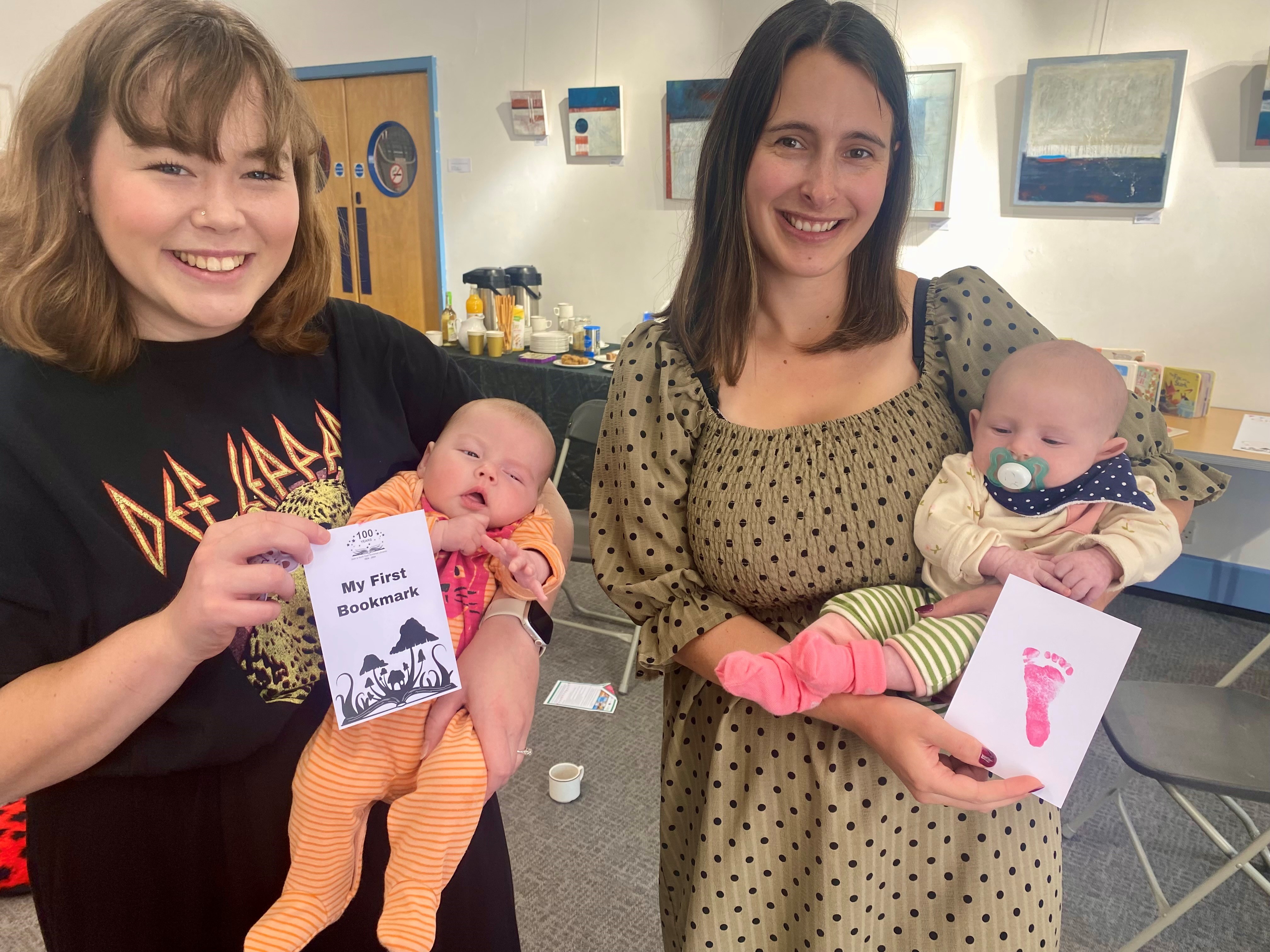 Two mothers hold their new babies in Bath Central Library, one holds My First Bookmark and the other a pink baby footprint on white paper