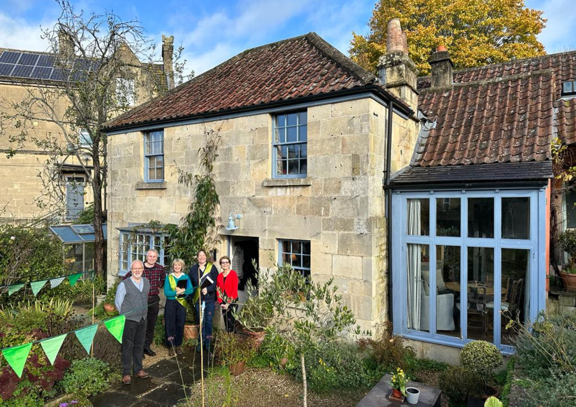 Five people stand at the entrance to a stone-built house with light blue window frames. Green bunting adorns the front garden.