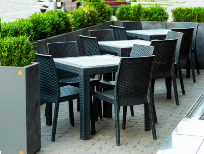 Grey weave tables and chairs on a street outside a restaurant. Planters surround the seating area.