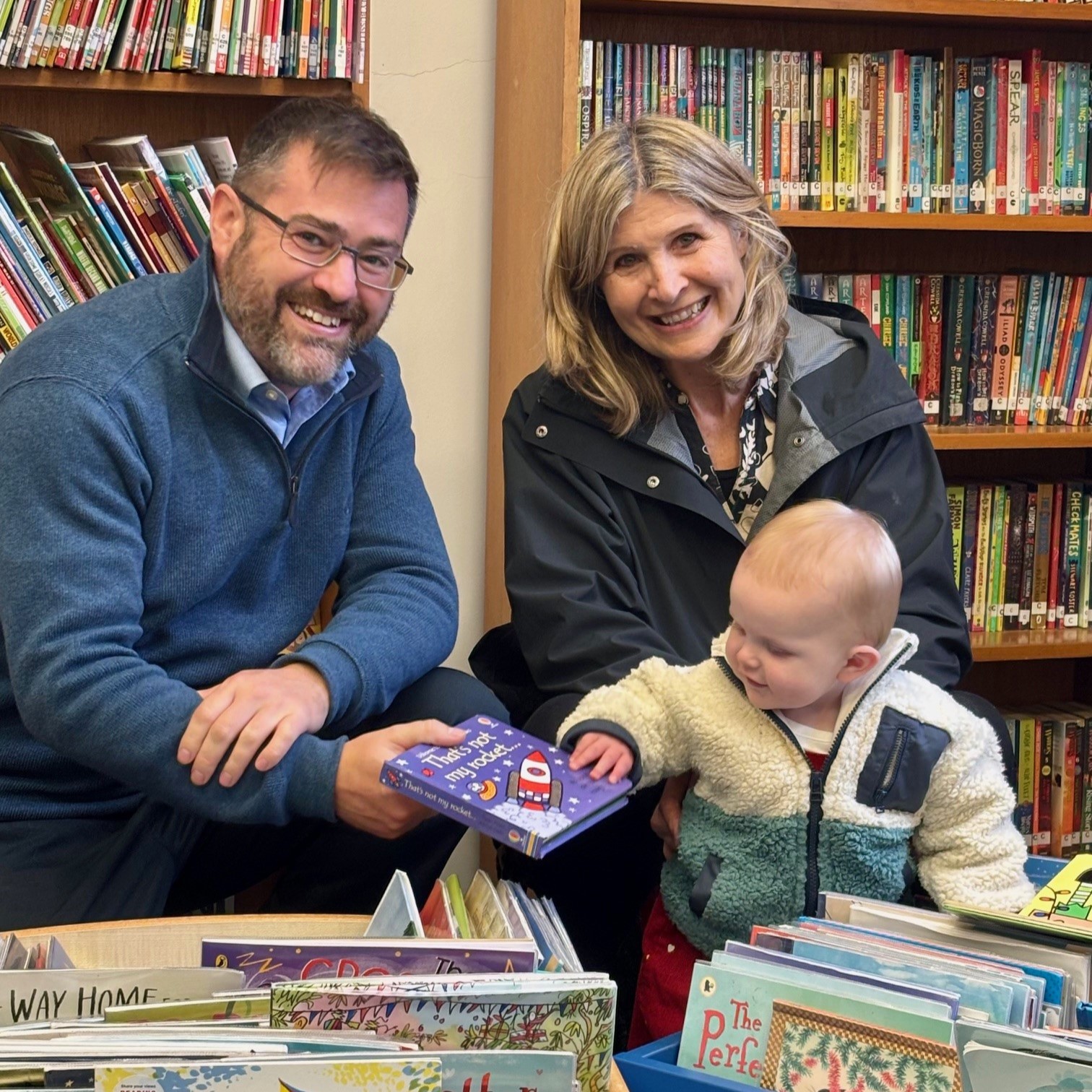 Two adults sit in a library with books on shelves behind them and in baskets in front of them. The person on the right holds a baby who is touching a children's book held by the person on the left