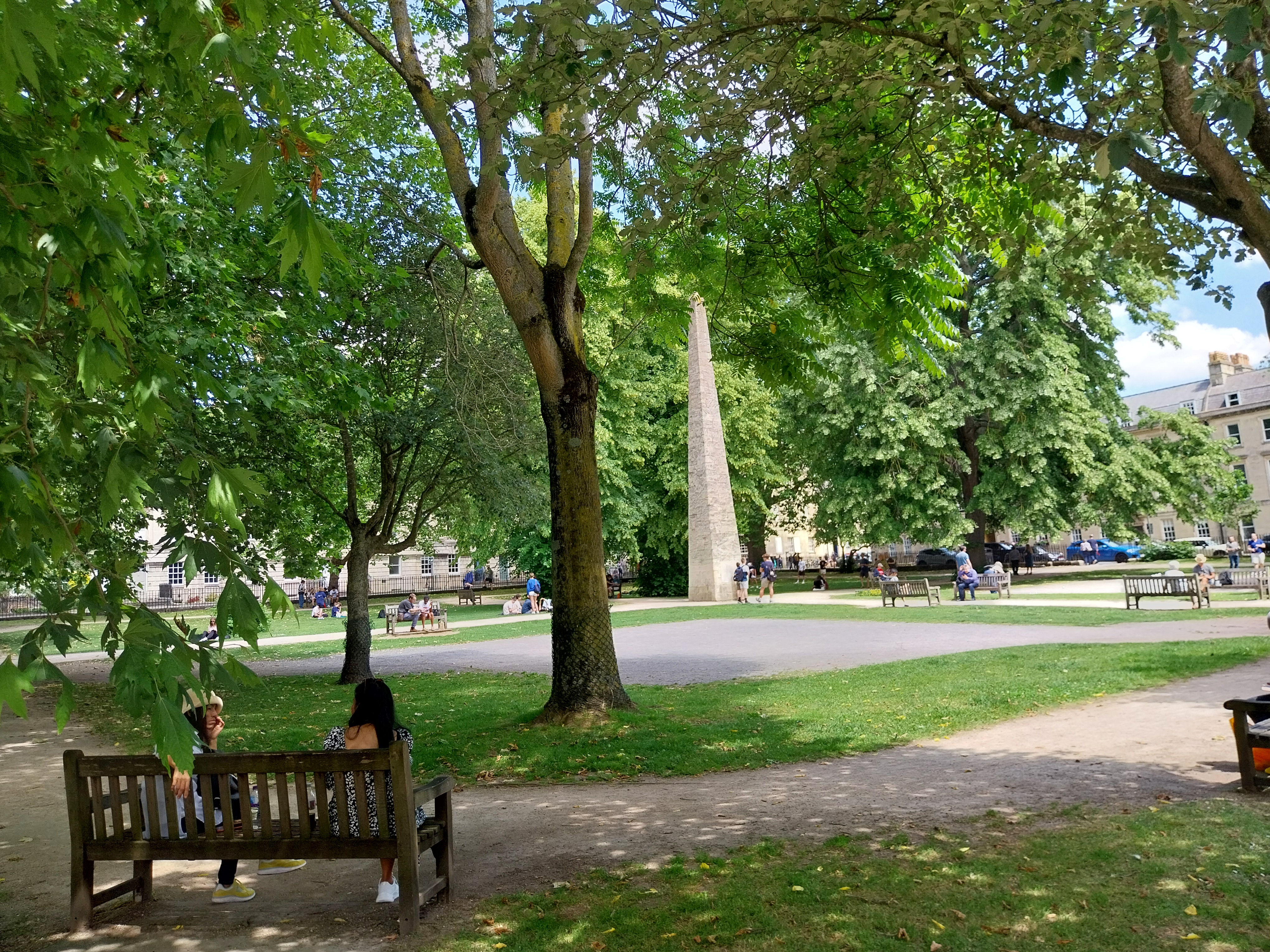 People sit on benches and stand amongst trees at Queen Square in Bath