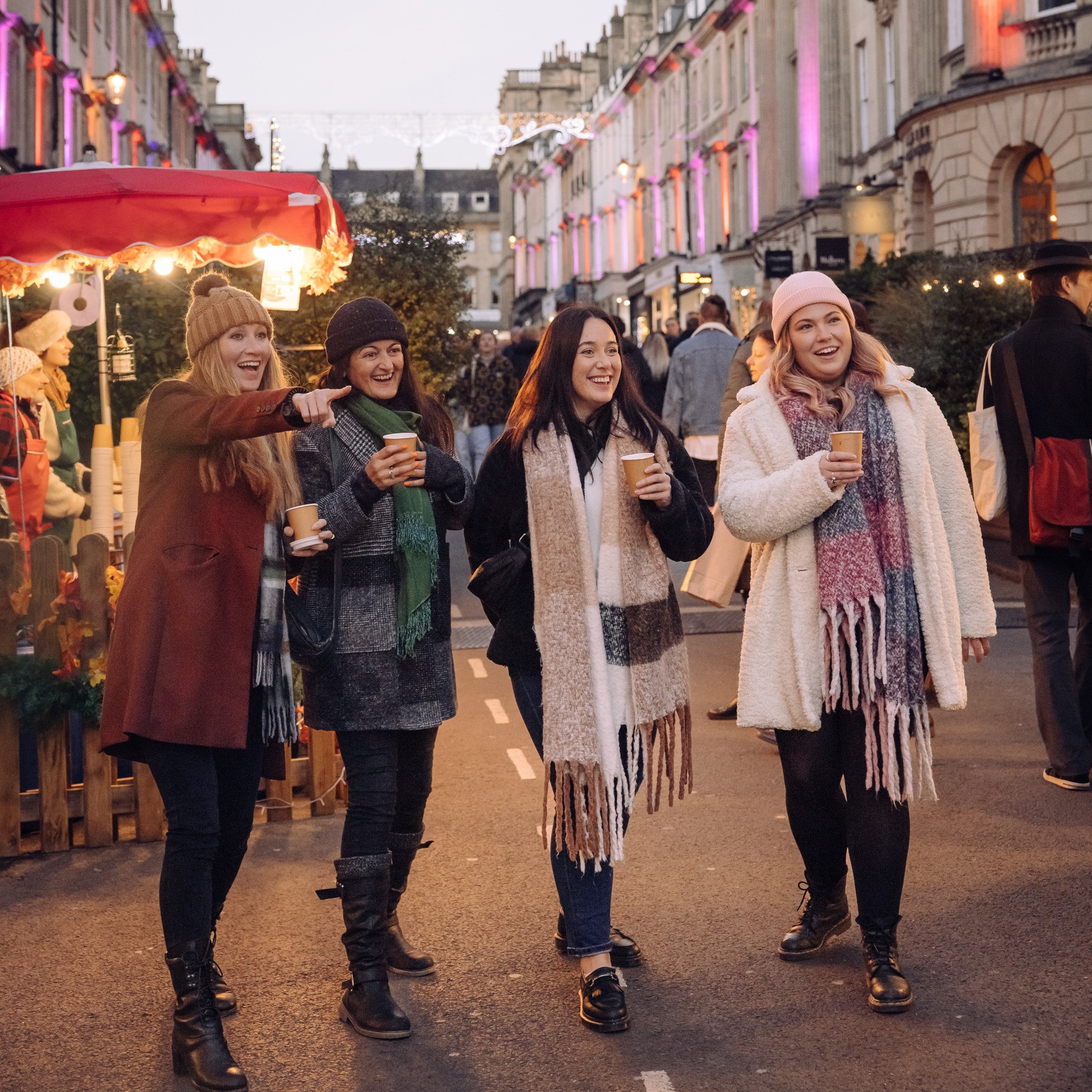 Four people stand in Milsom Street with Christmas market chalets behind, they all hold cups and one of the people is pointing