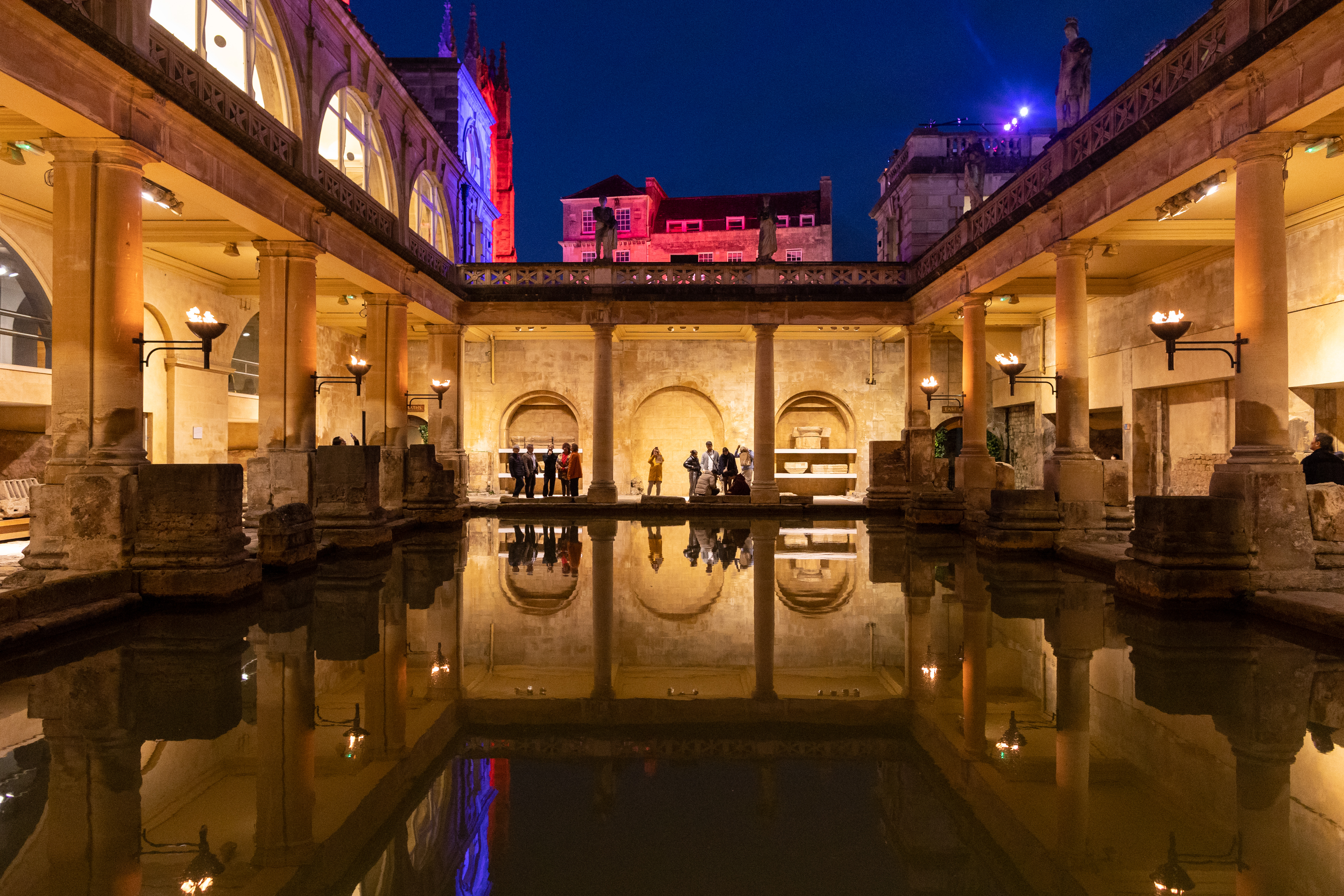 The Great Bath at the Roman Baths at dusk, with surrounding buildings lit up in coloured lights