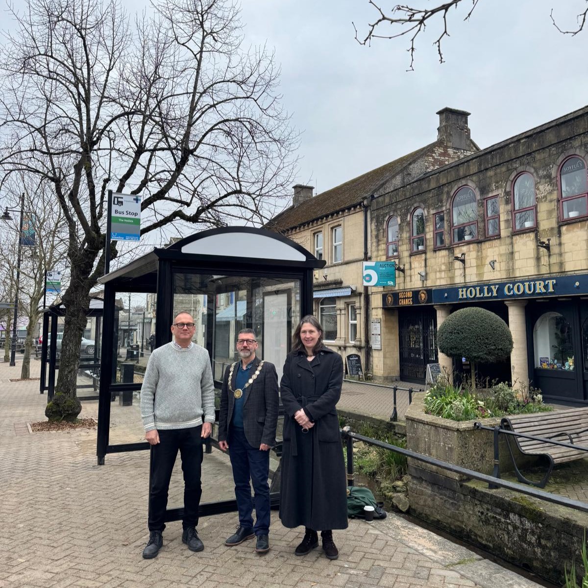 Three people stand at a bus stop in Midsomer Norton