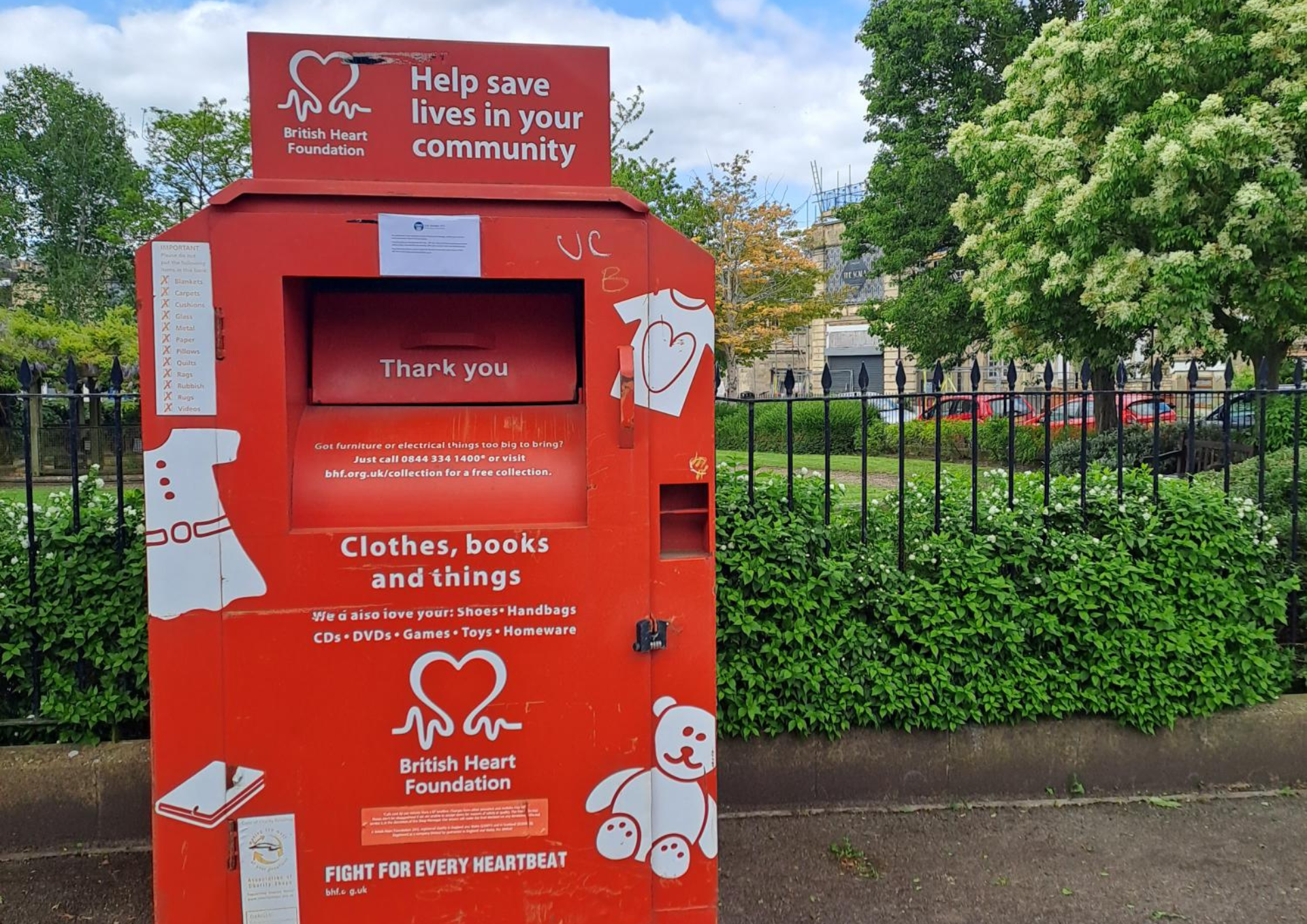 a photo of a red British Heart Foundation donation bank in front of a green area of park