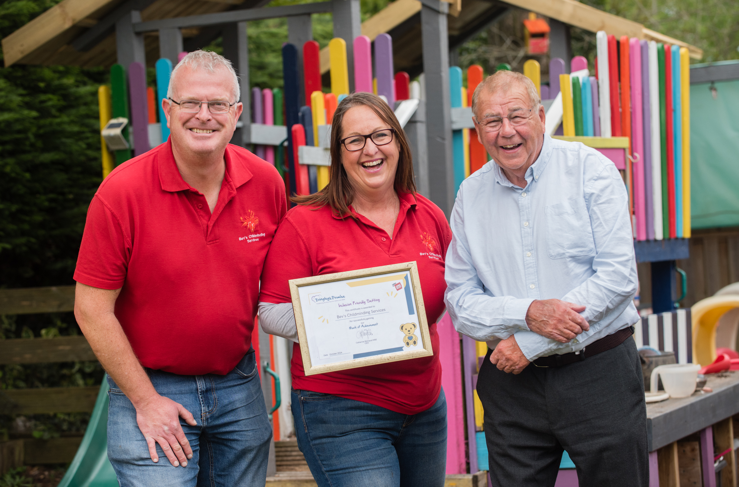 Three people stand in front of play equipment in a garden, one holds a framed certificate