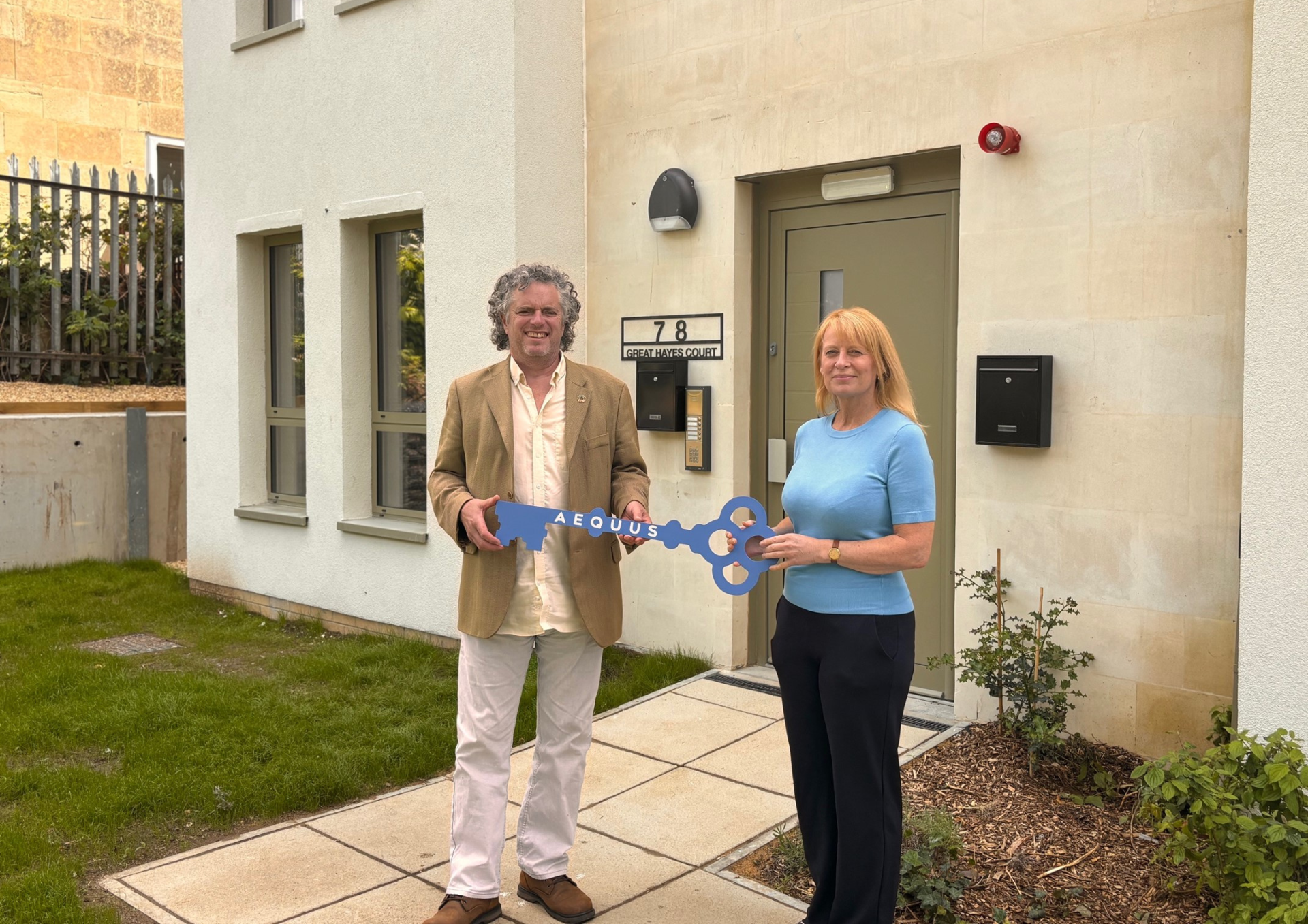 a photo of a man and woman outside a new housing development. They are holding a blue cut out of a key with the word Aequus written on it. it