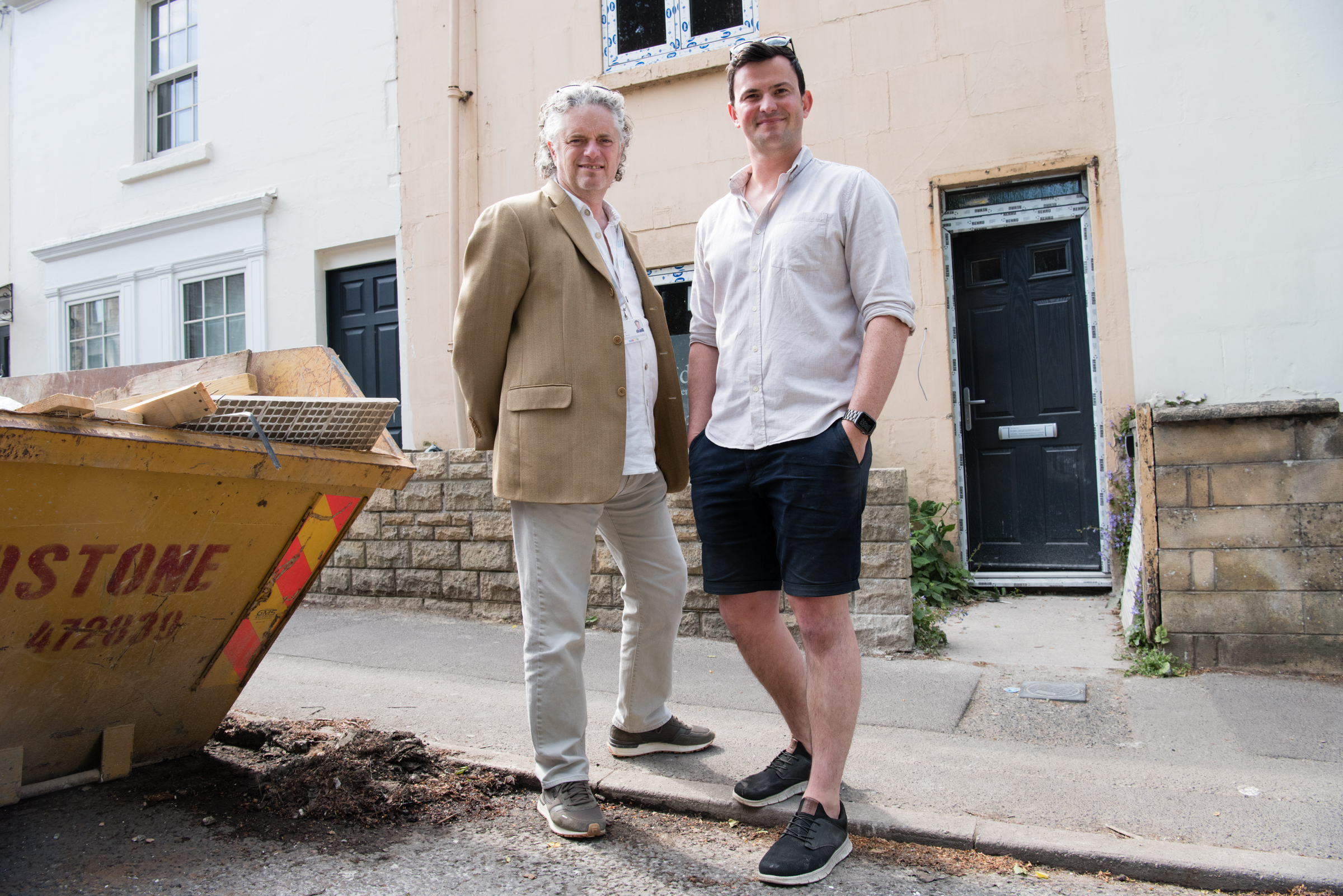 Two people stand in front of a house where works are taking place with a skip on the left of the photo