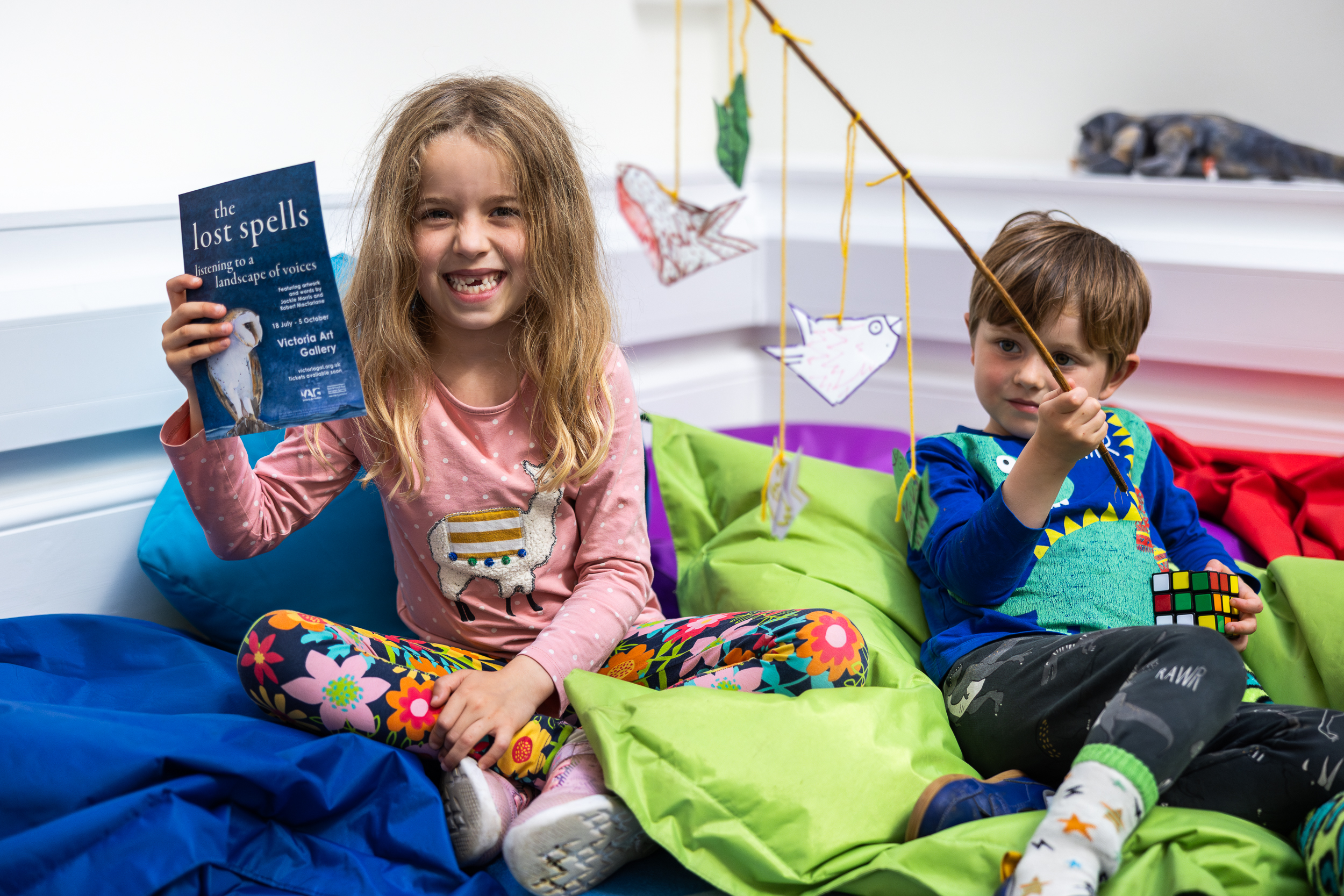 Two young children sitting on beanbags. The girl is holding a flyer for The Lost Spells exhibition, the boy is holding a stick with handmade birds hanging from it.