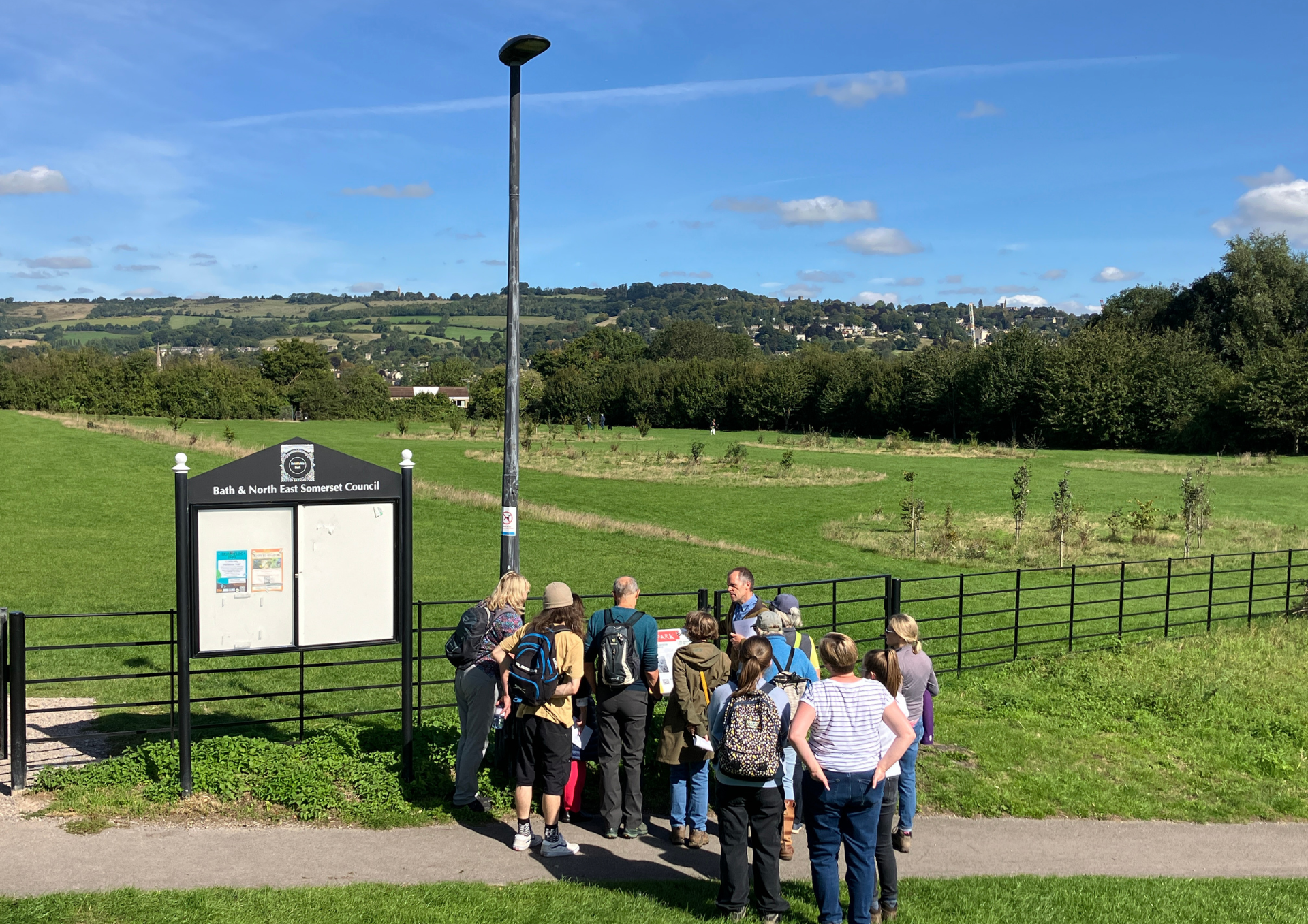 a photo of walkers grouped around a leader. they are on a footpath outside a park with grass in front and trees in the background