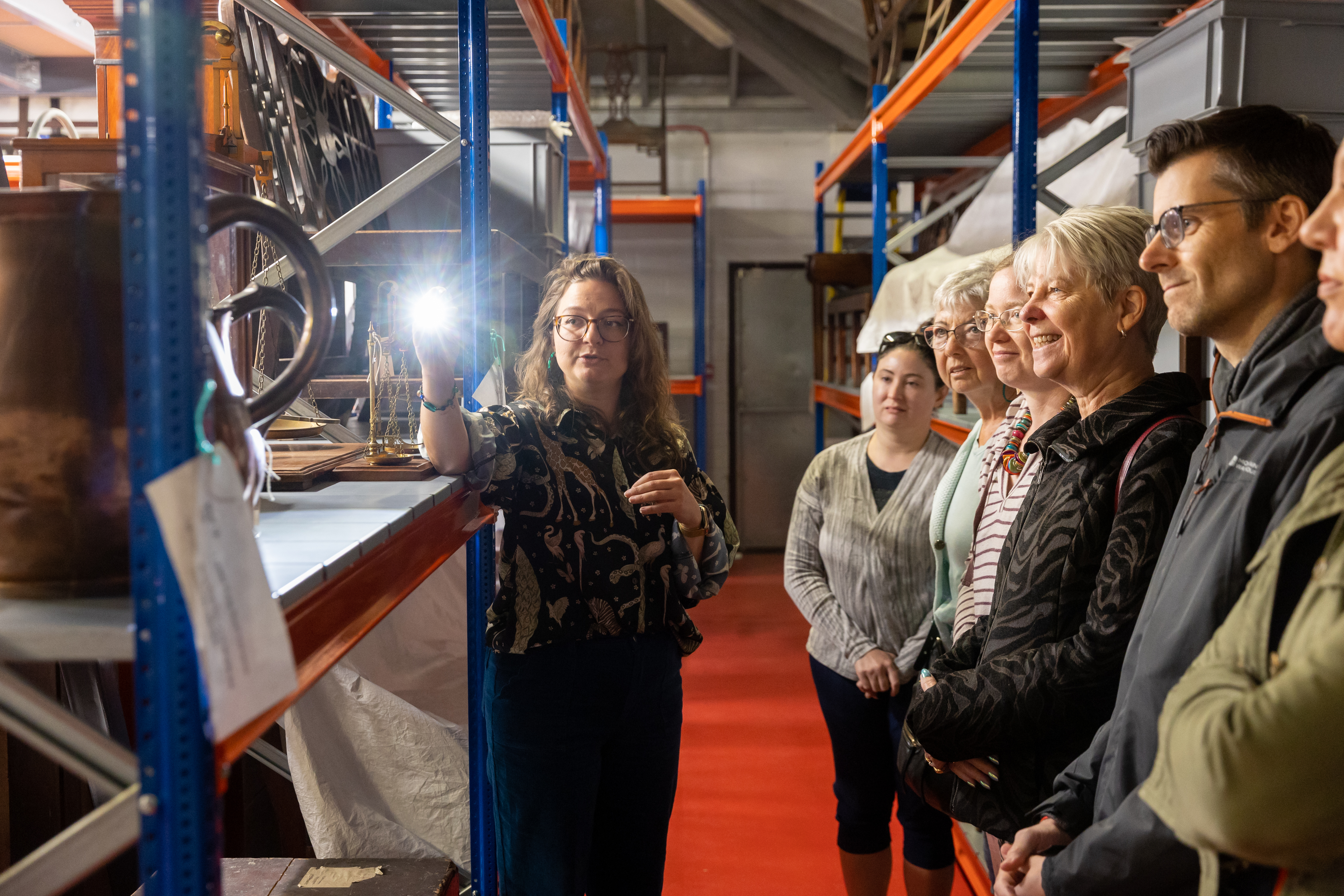 A small group of people in a store room, watching a curator shine a light on the objects on a shelf