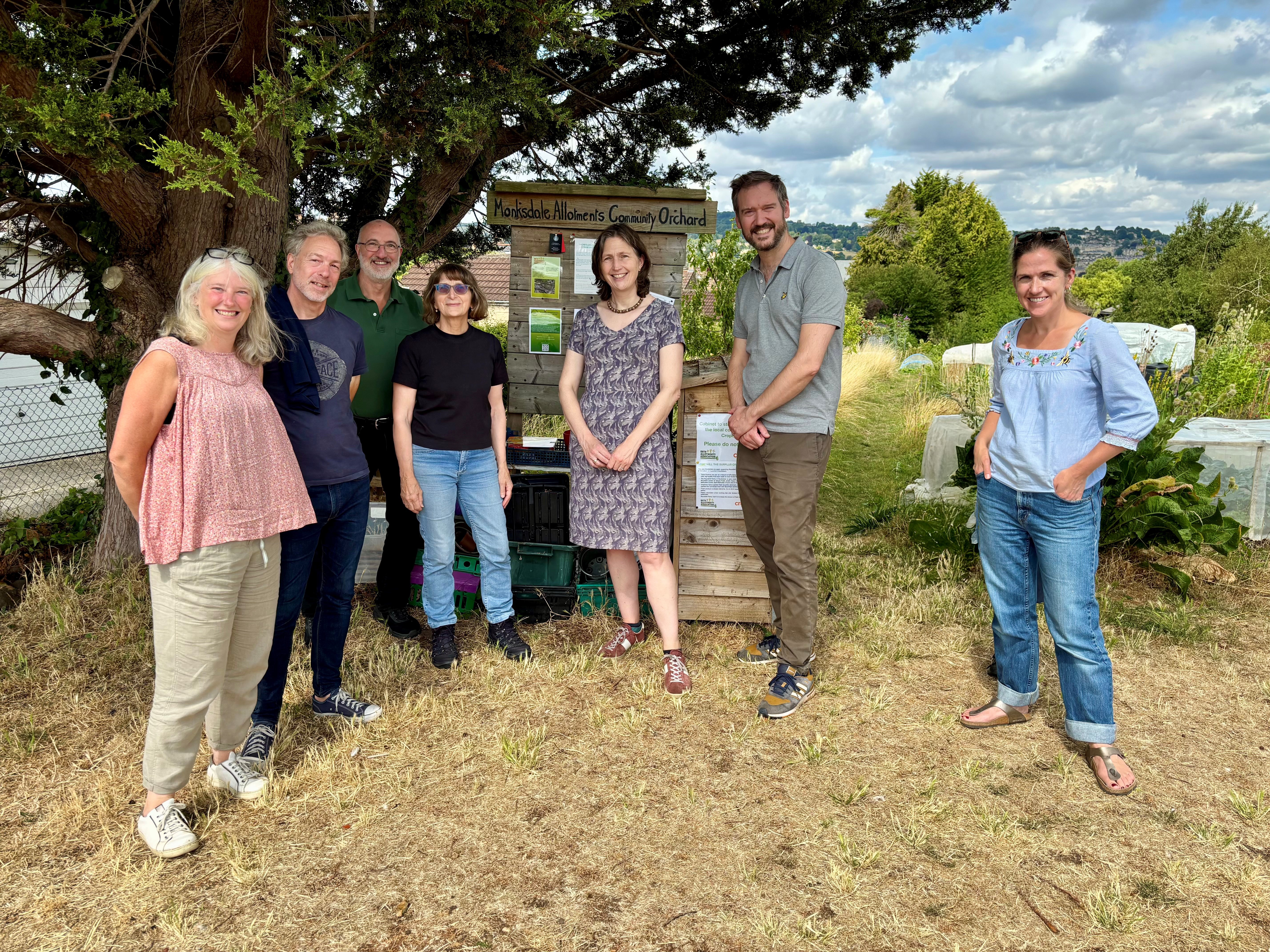 A group of men and women standing in front of an allotment with a tree behind them