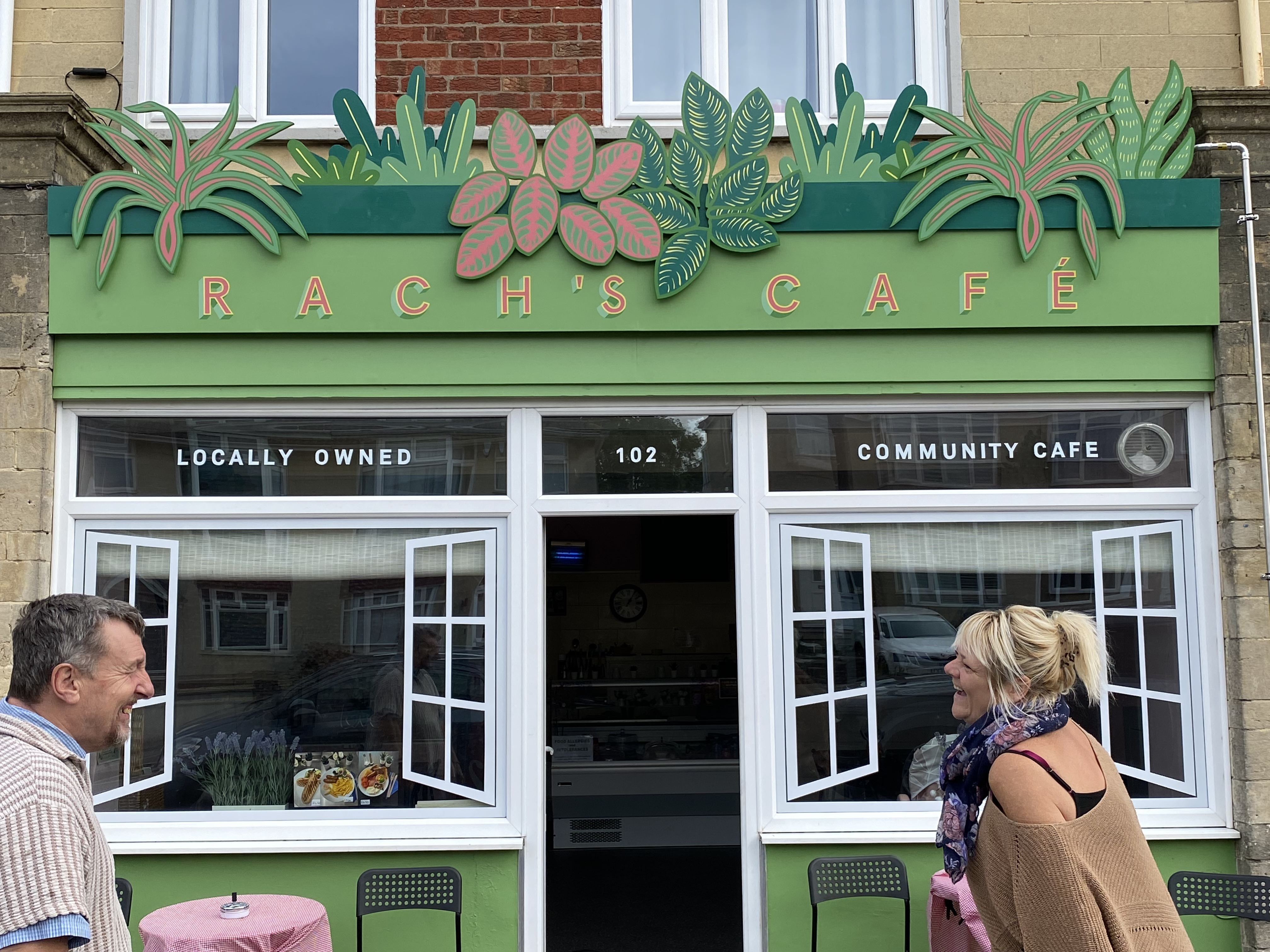 Two people stand outside a cafe, they face each other and laugh