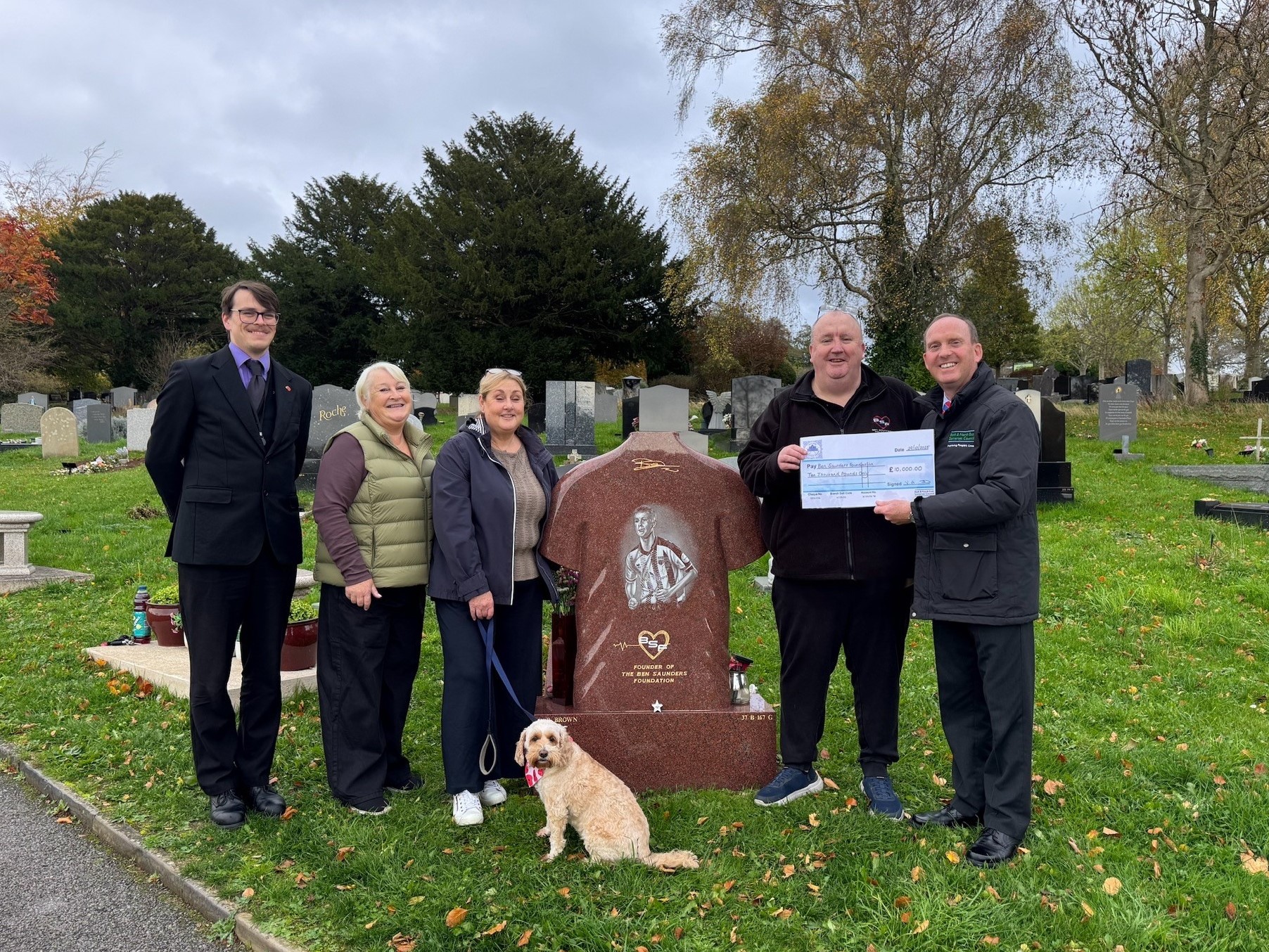 A family and cemetery staff stand with a dog by a grave, one person holds a large cheque