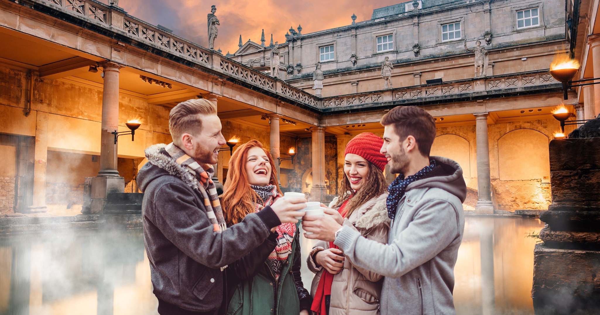 Four people making a toast with drinks in paper cups in front of the torchlit Great Bath