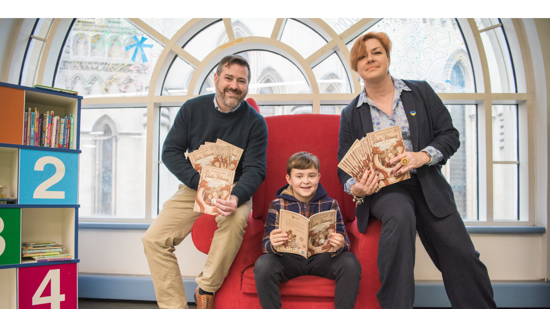 A child sits on a large red armchair in a library holding books, an adult sits on each arm of the chair also holding books.