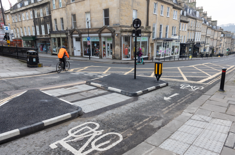 A person cycles at a junction wearing a bright orange hi vis jacket