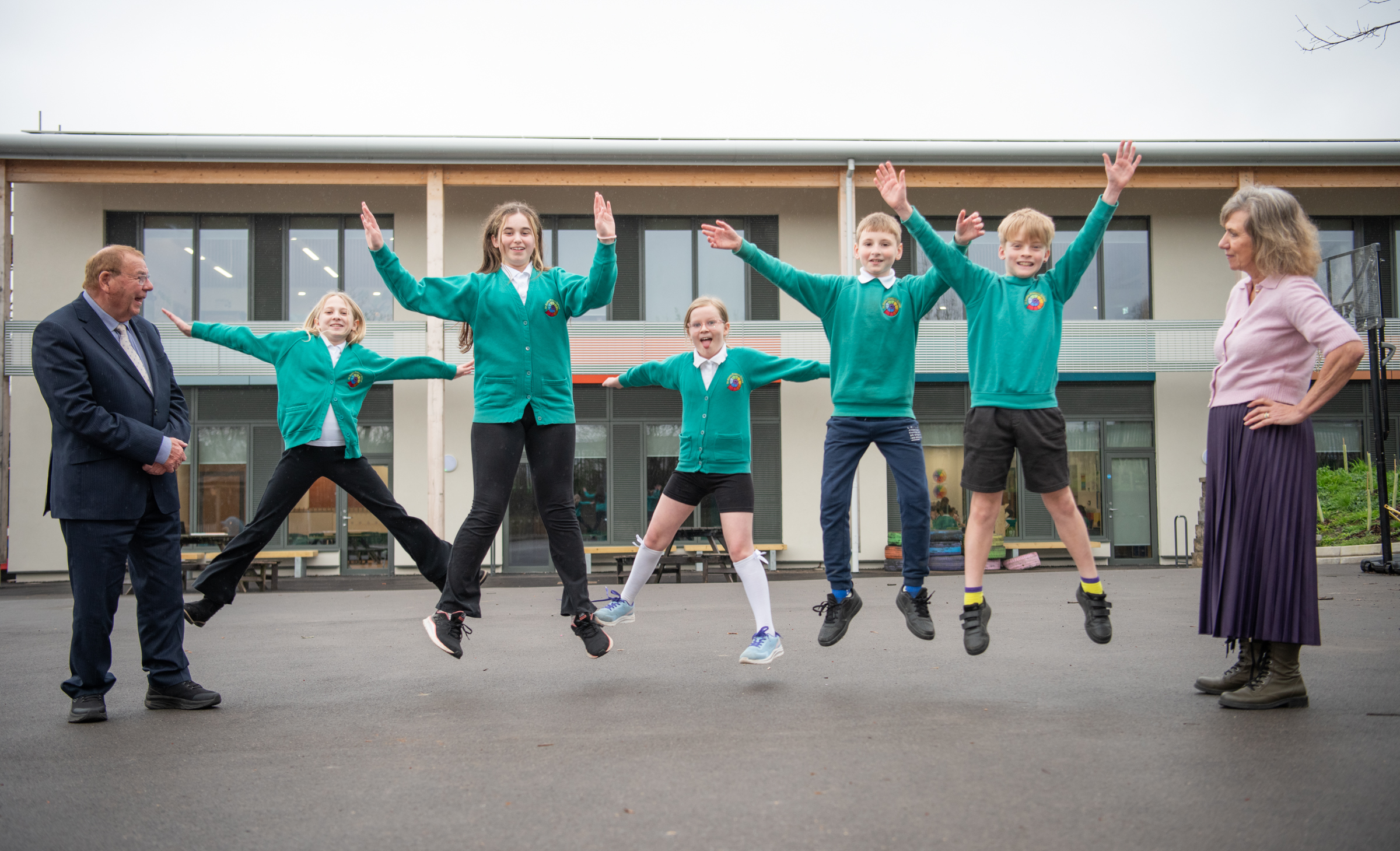 Five children jump in the air at a school playground as adults look on