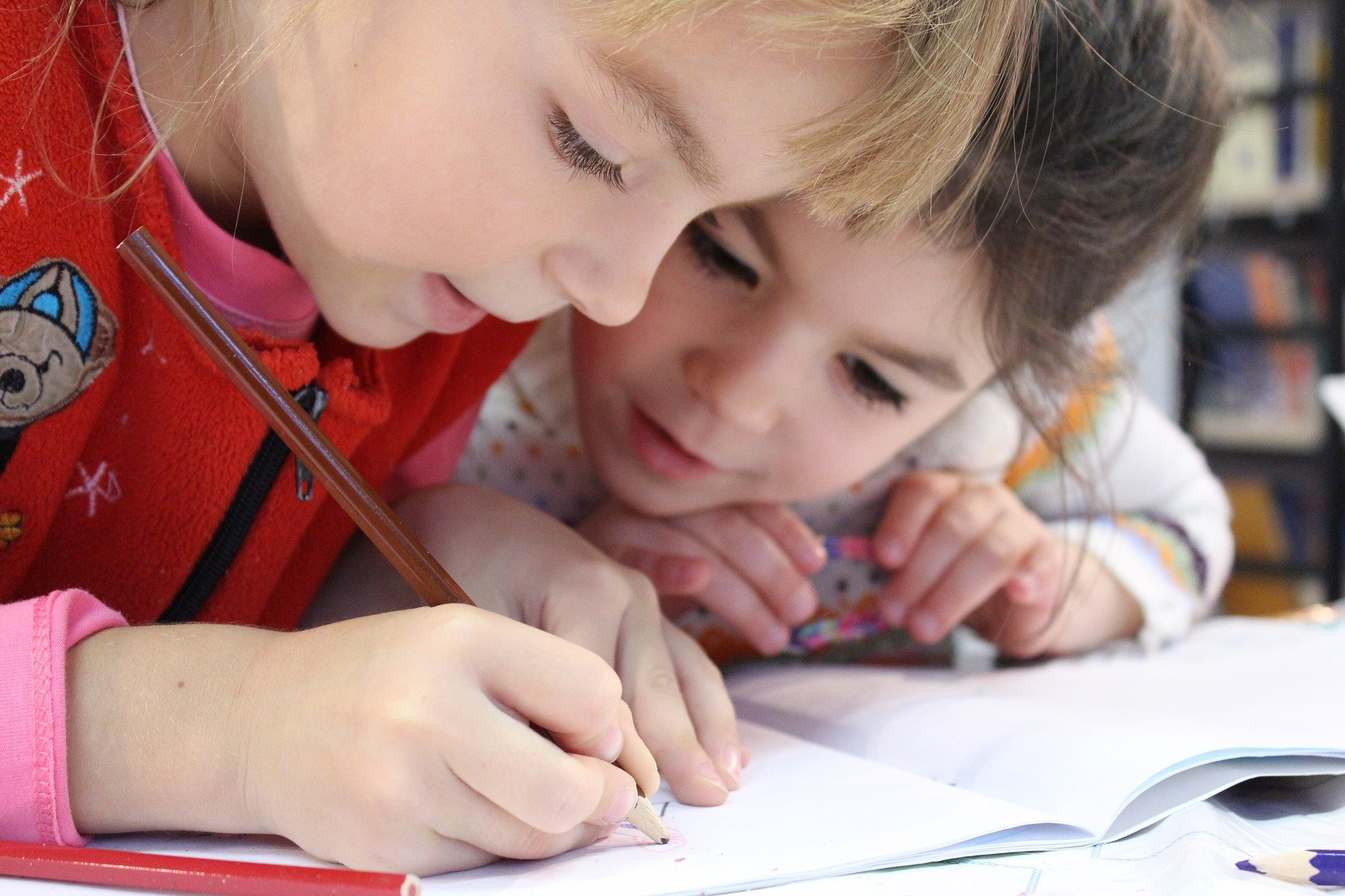 Two children look at a book, one holds a pencil and writes