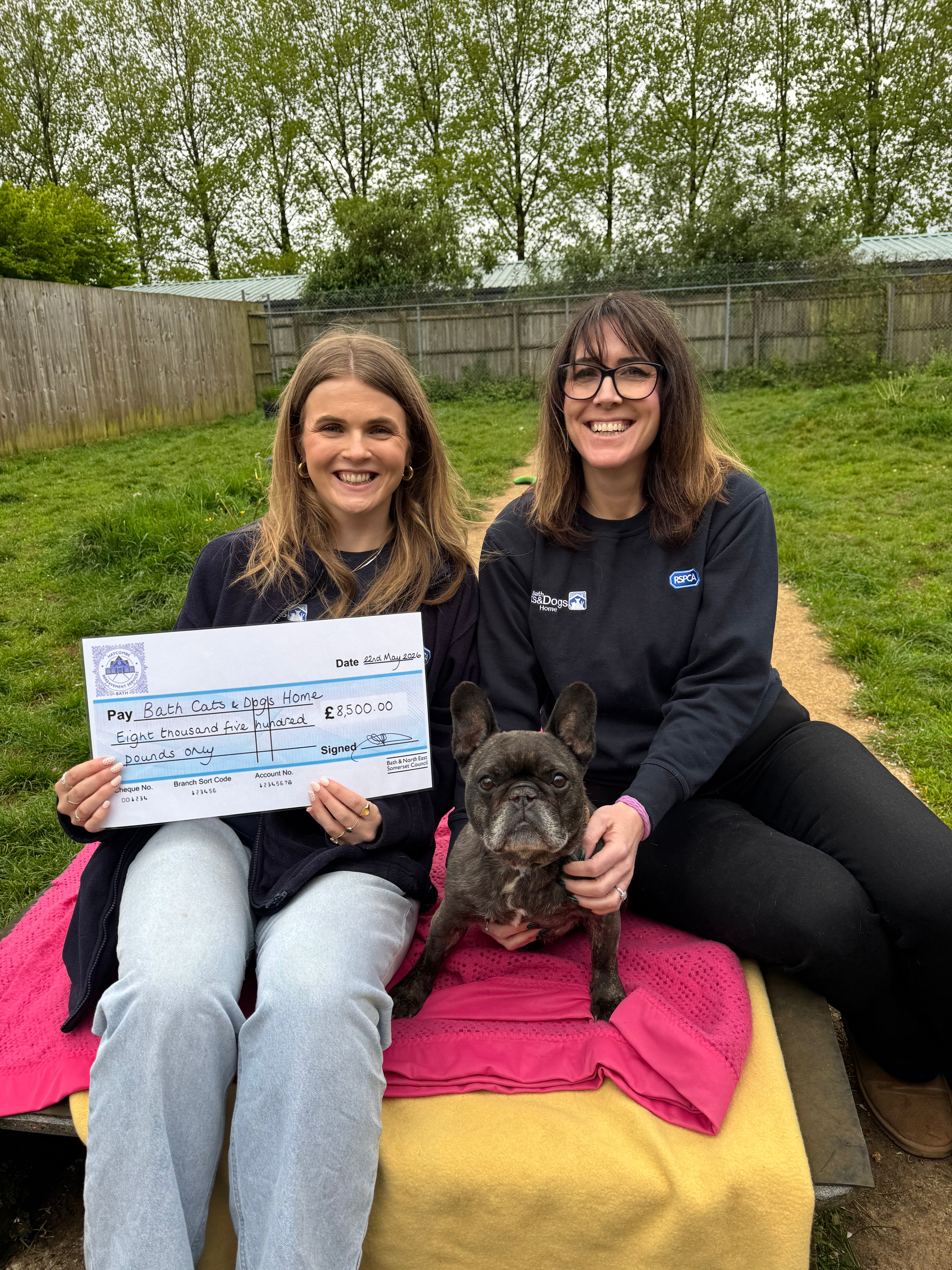 Two people hold a large cheque with a dog sitting in front of them
