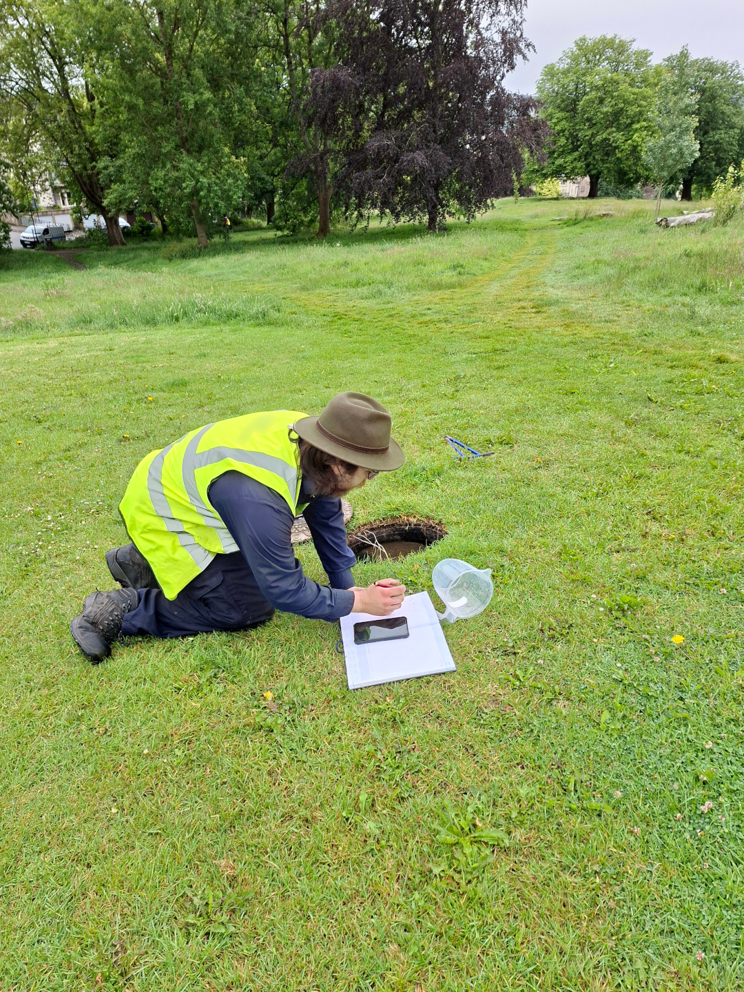 Person crouches on the ground taking notes near a hole in the ground in Royal Victoria Park