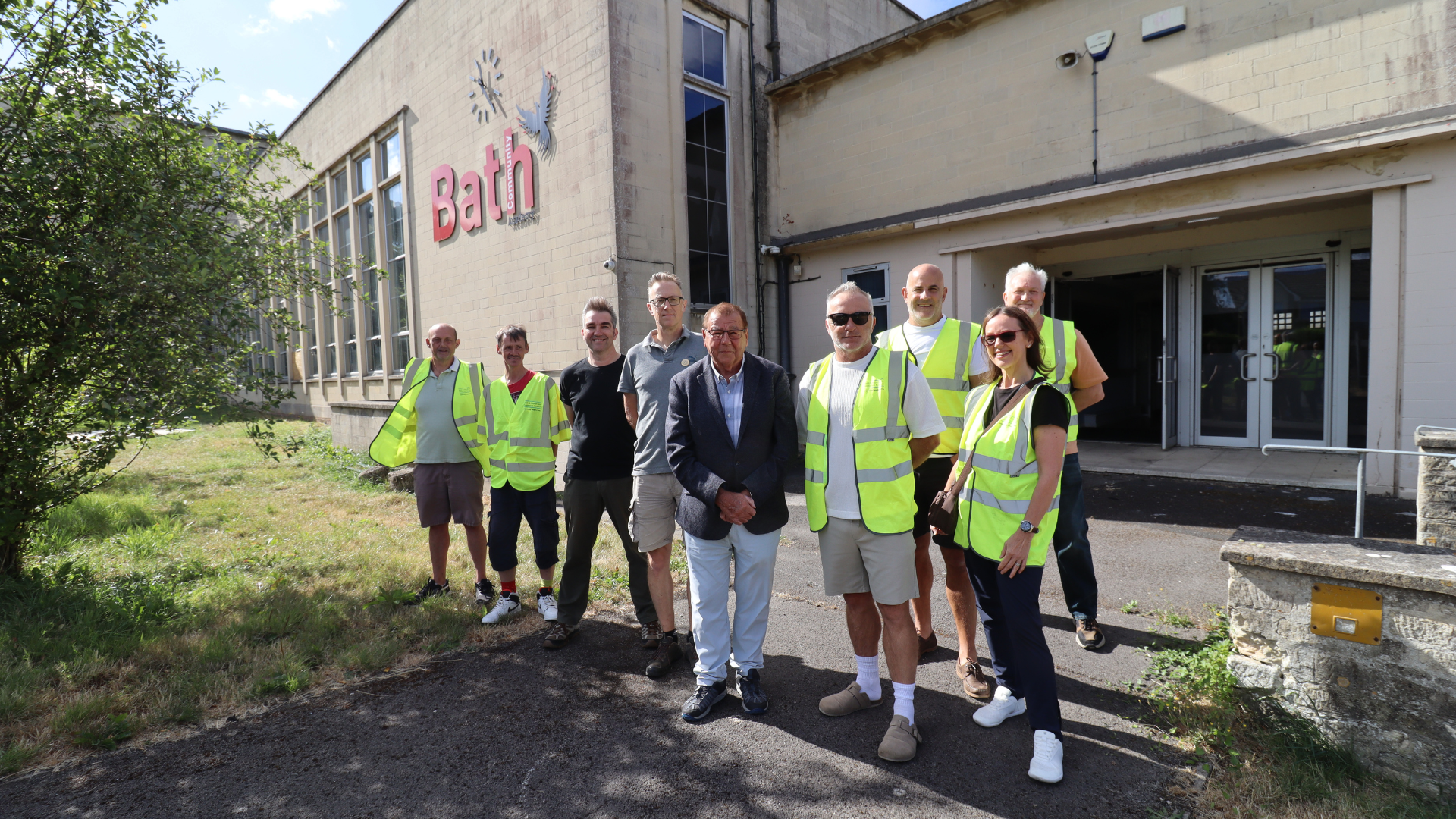 a group of people standing in front of an old school building. They have high visibility jackets on. The sign on the wall says Bath and there is an image of a bird next to it and a clock.