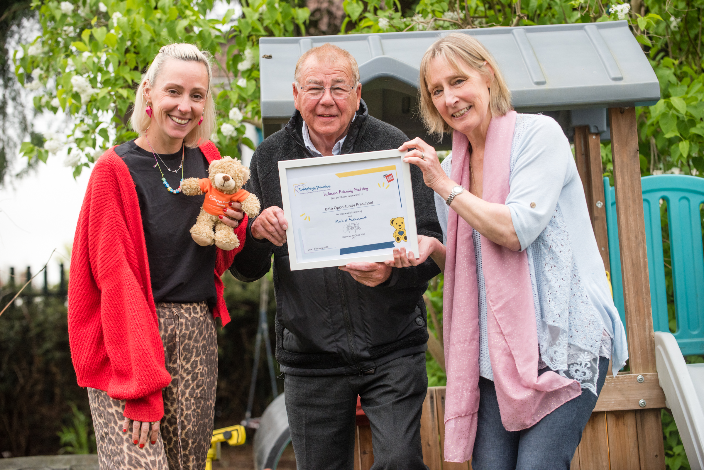 Three people stand in front of play equipment in a garden, one holds a framed certificate