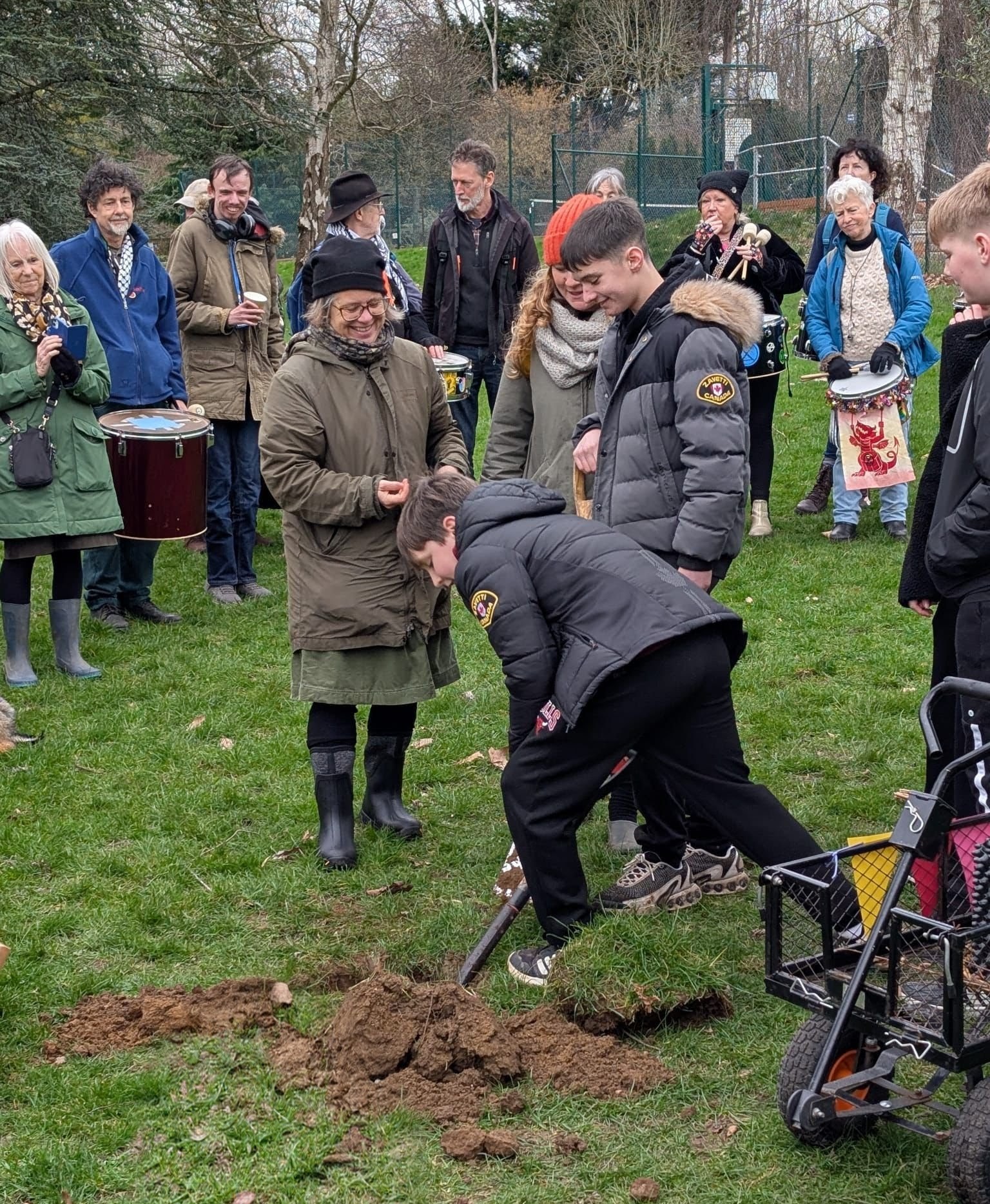 A young person digs with a spade to plant a sapling as a crowd look on